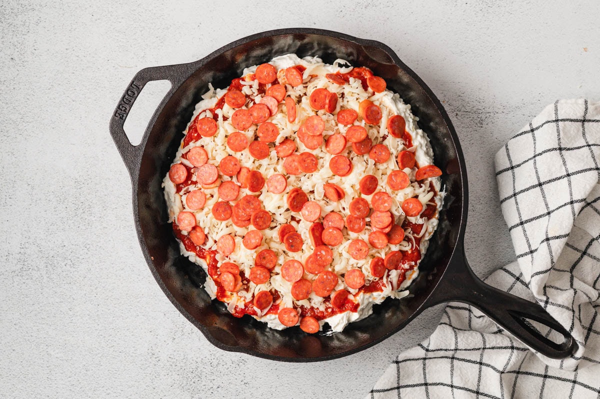 Unbaked pizza dip with cheese and mini pepperoni on top all in a cast iron pan.