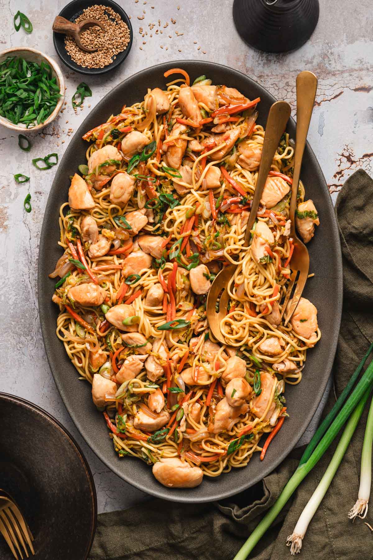 Chicken chow mein on a black platter with gold utensils