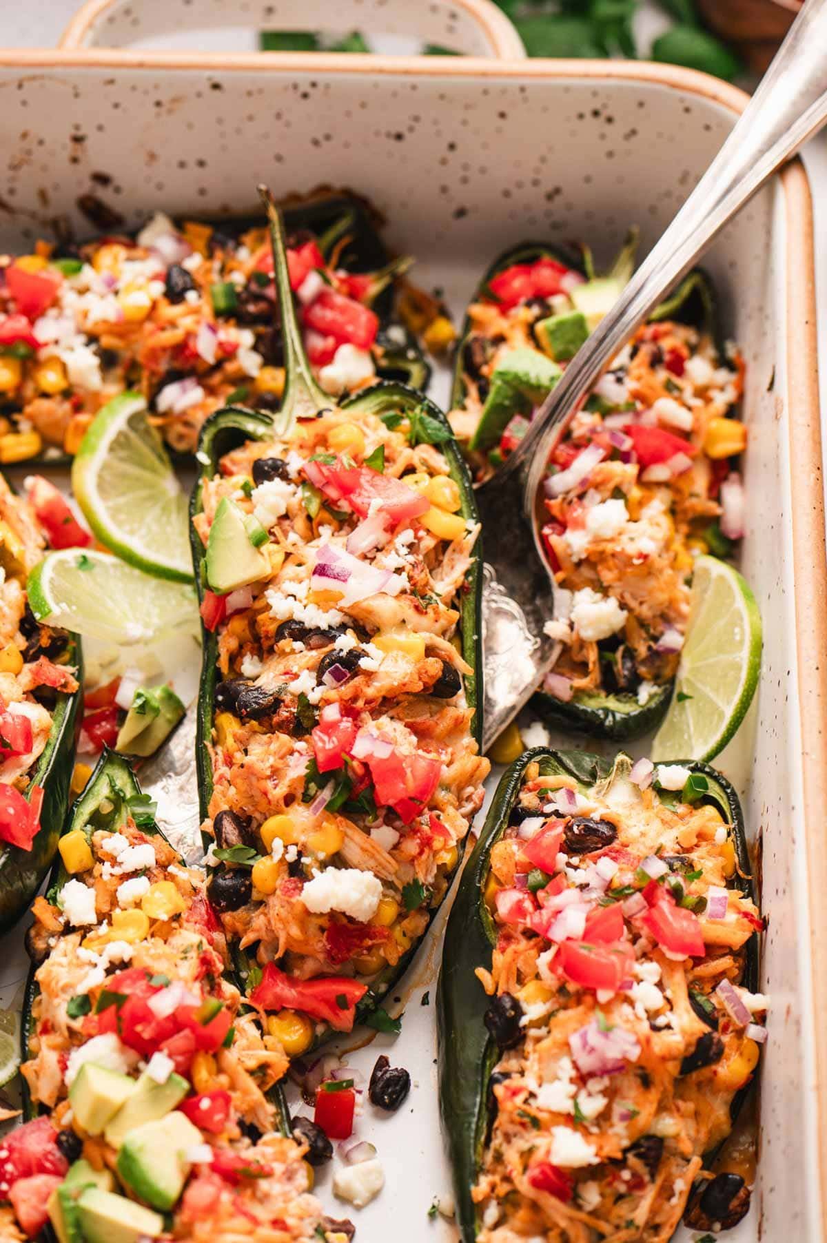 Close up of a chicken, bean, and corn stuffed poblano pepper with garnishes like lime wedges and avocado, all in a baking dish.