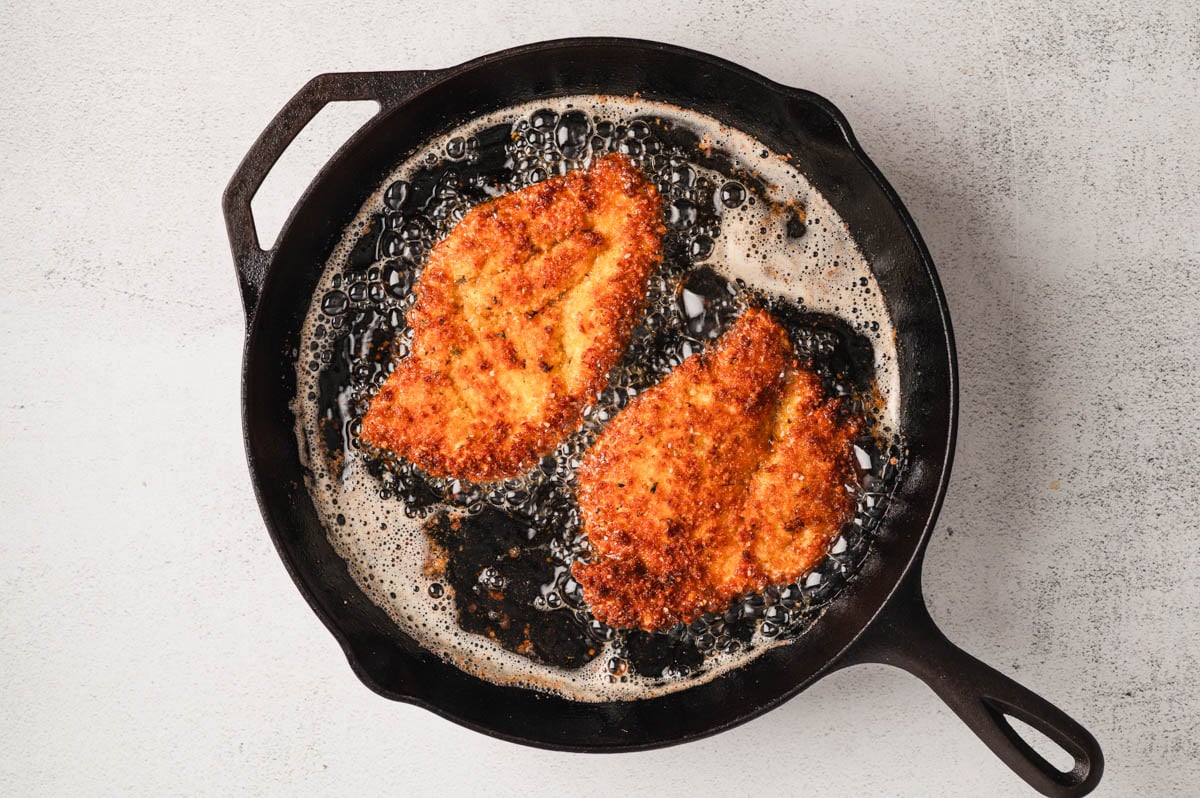 Two breaded chicken cutlets frying in a cast iron pan.