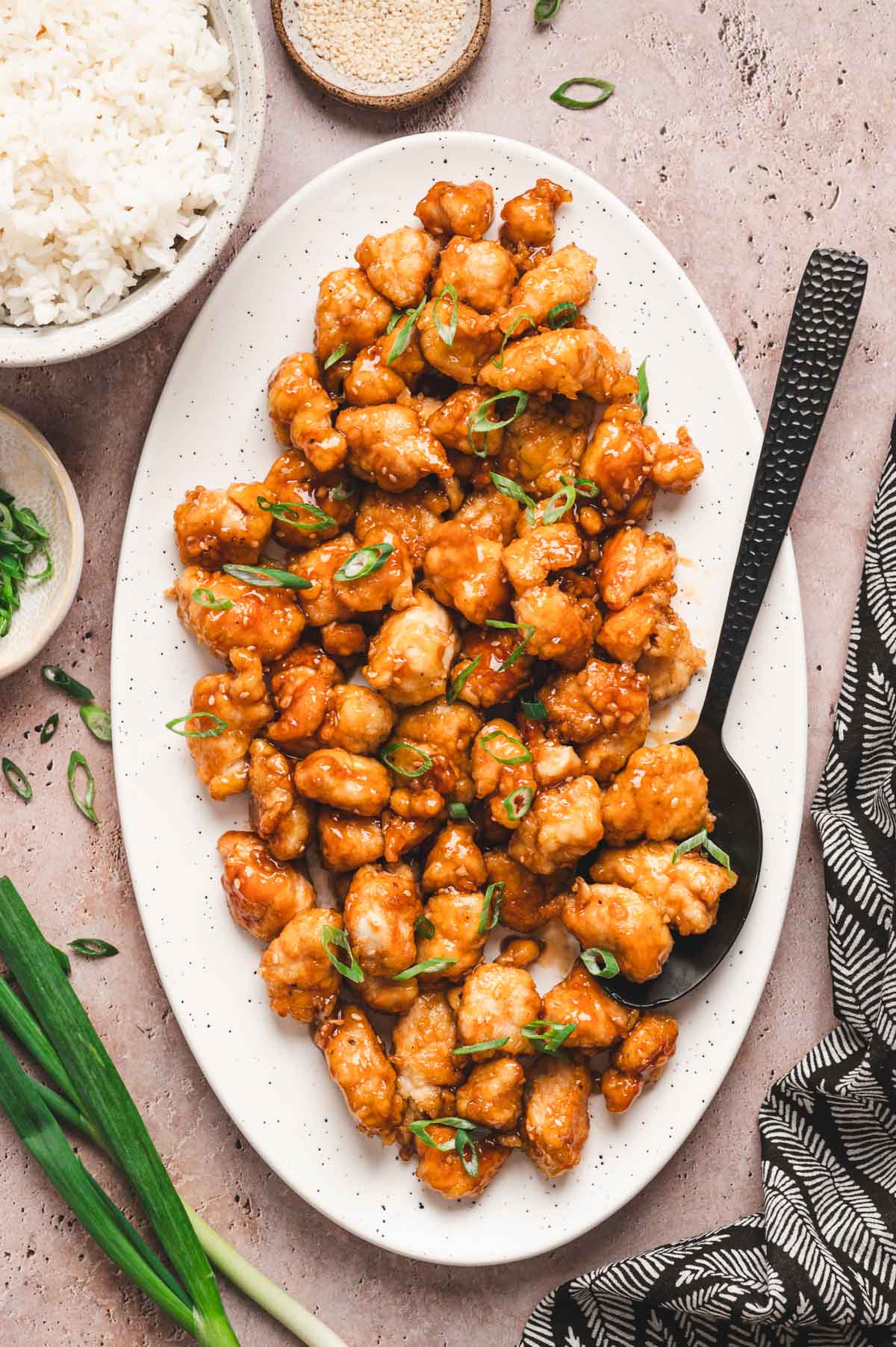 Sesame chicken with green onions on top all on a white platter with black serving spoon.