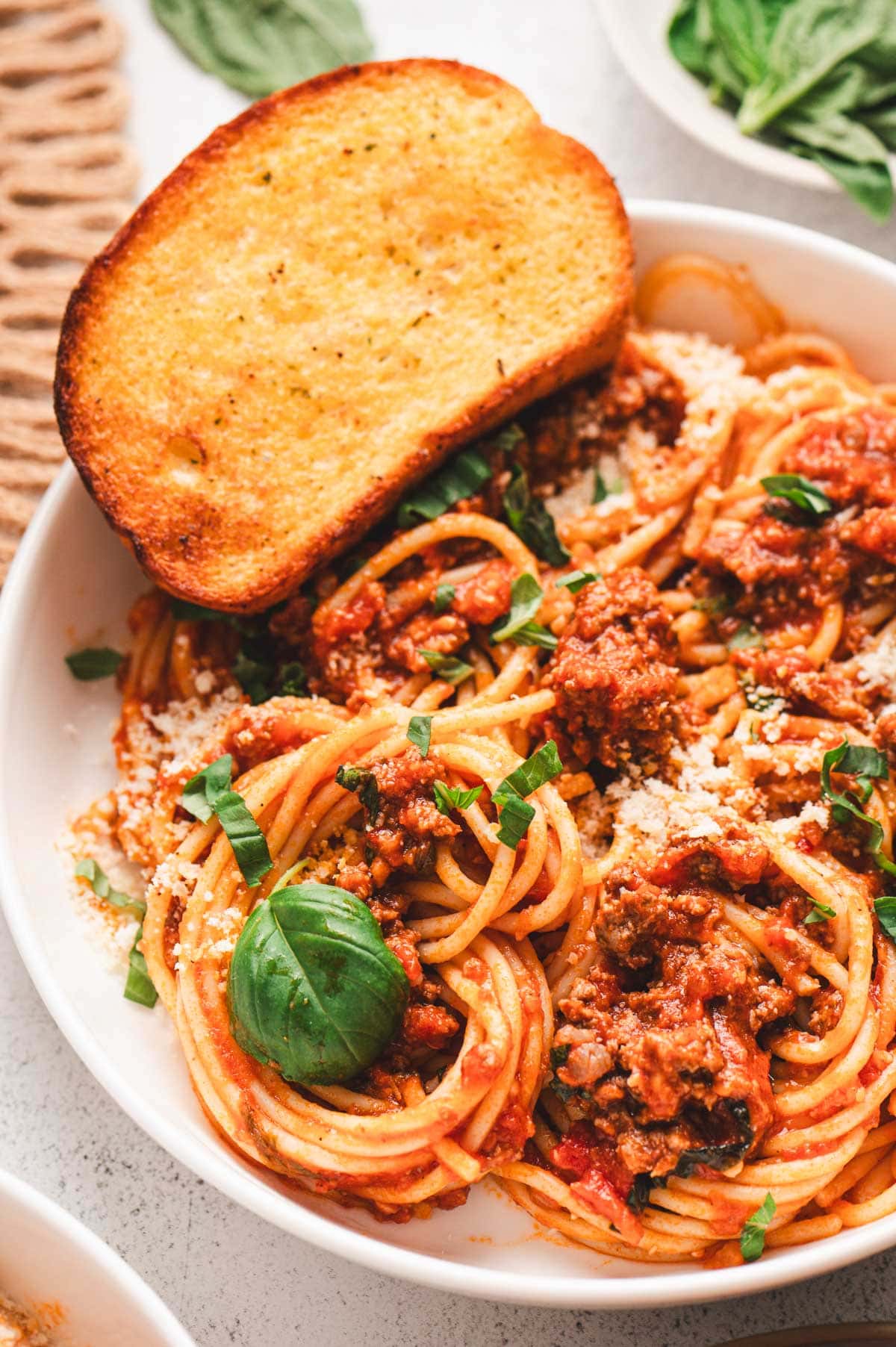 Spaghetti with meat sauce in a white serving dish with fresh basil and a piece of garlic bread on top.