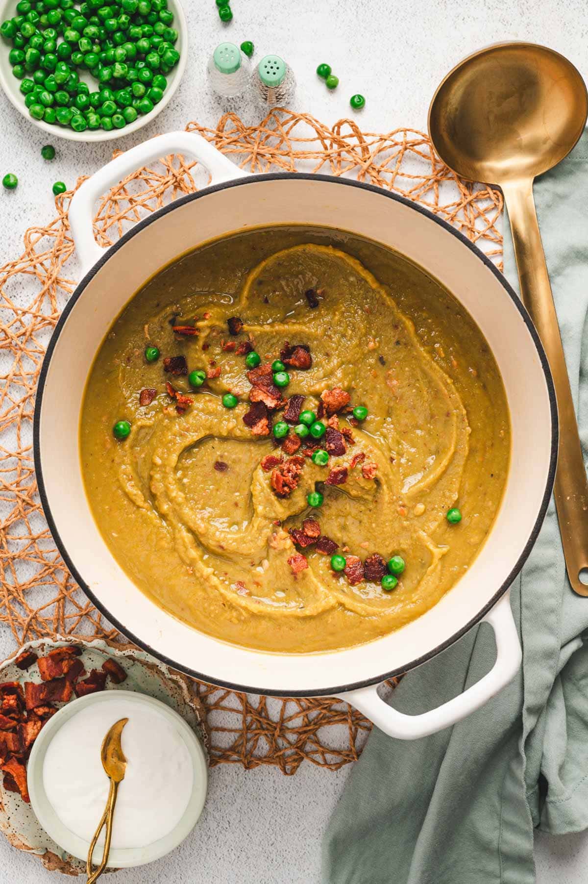 This Split Pea Soup Recipe with Bacon has a thick and creamy, split pea base, loaded with veggies, seasonings and tons of crunchy, salty bacon. Split pea soup with garnish of peas and bacon in a white enameled pot with gold ladle