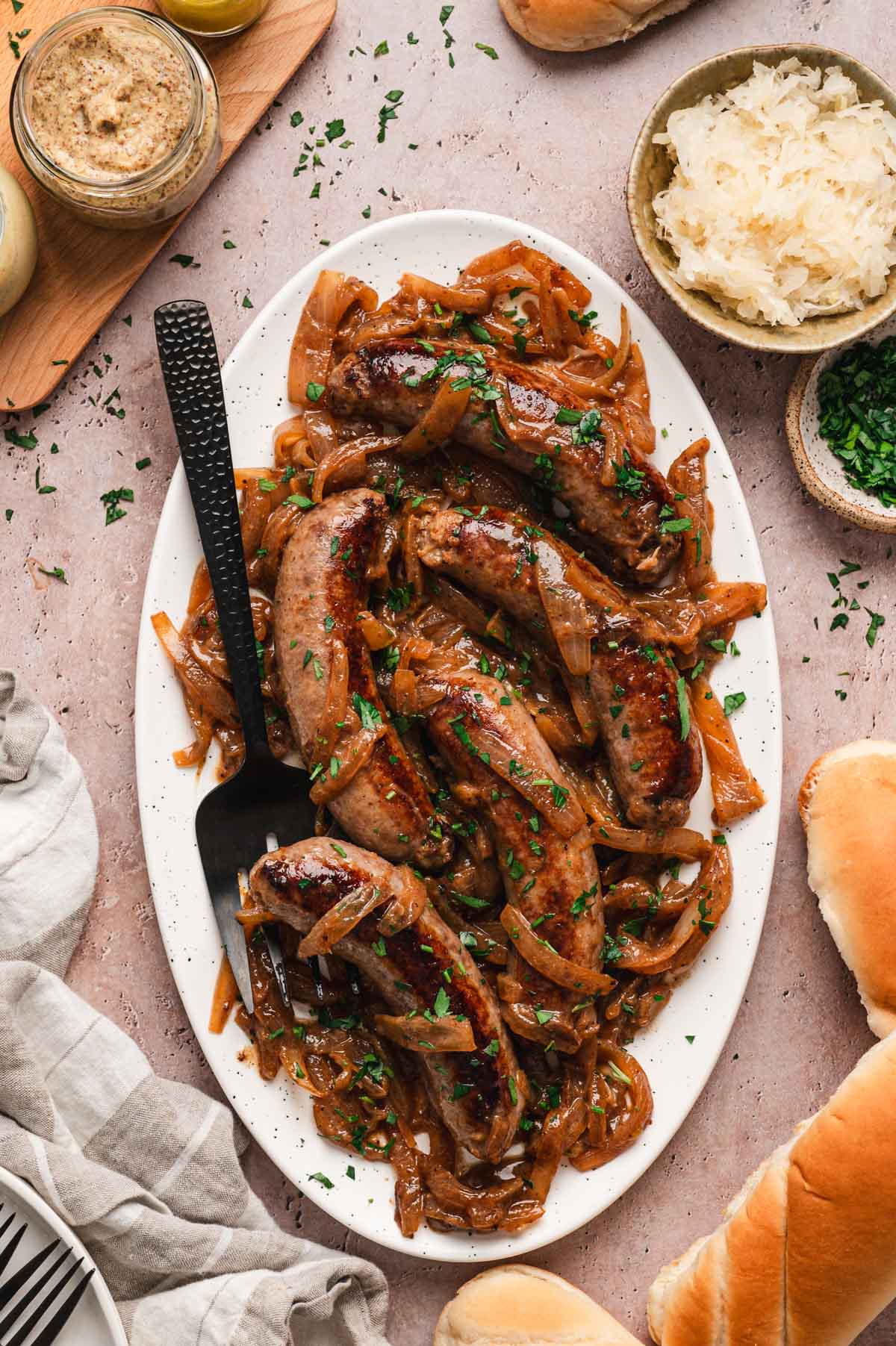 Bratwurst sausages with onions on a white serving platter with black serving utensil.