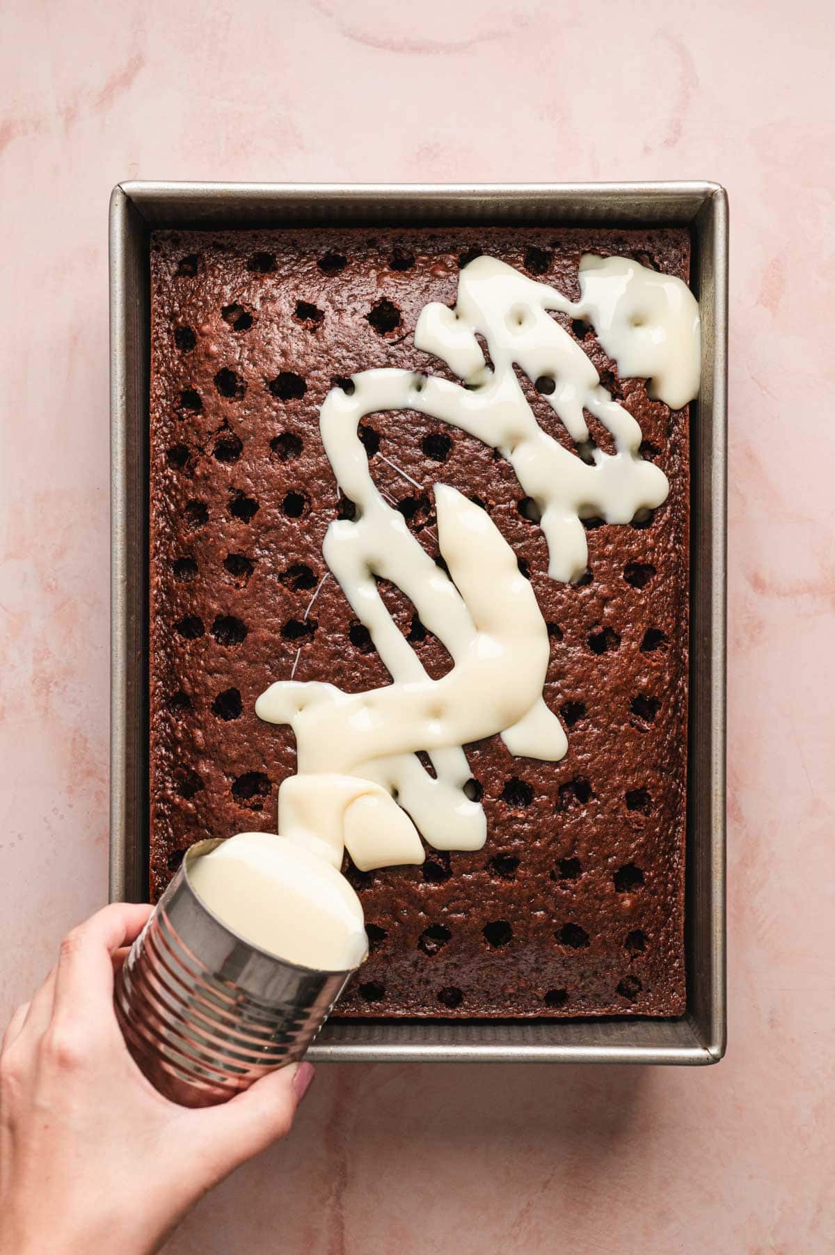 Sweetened condensed milk being poured over chocolate cake.