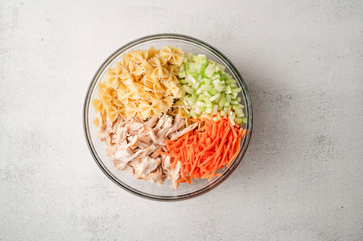 Pasta, celery, shredded chicken, and matchstick carrots in a large glass bowl.