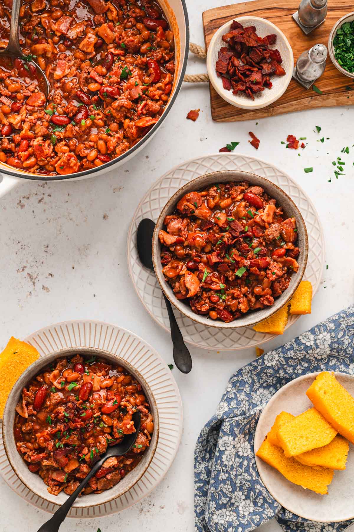 Bowls of meaty baked beans next to cornbread pieces on a plate.