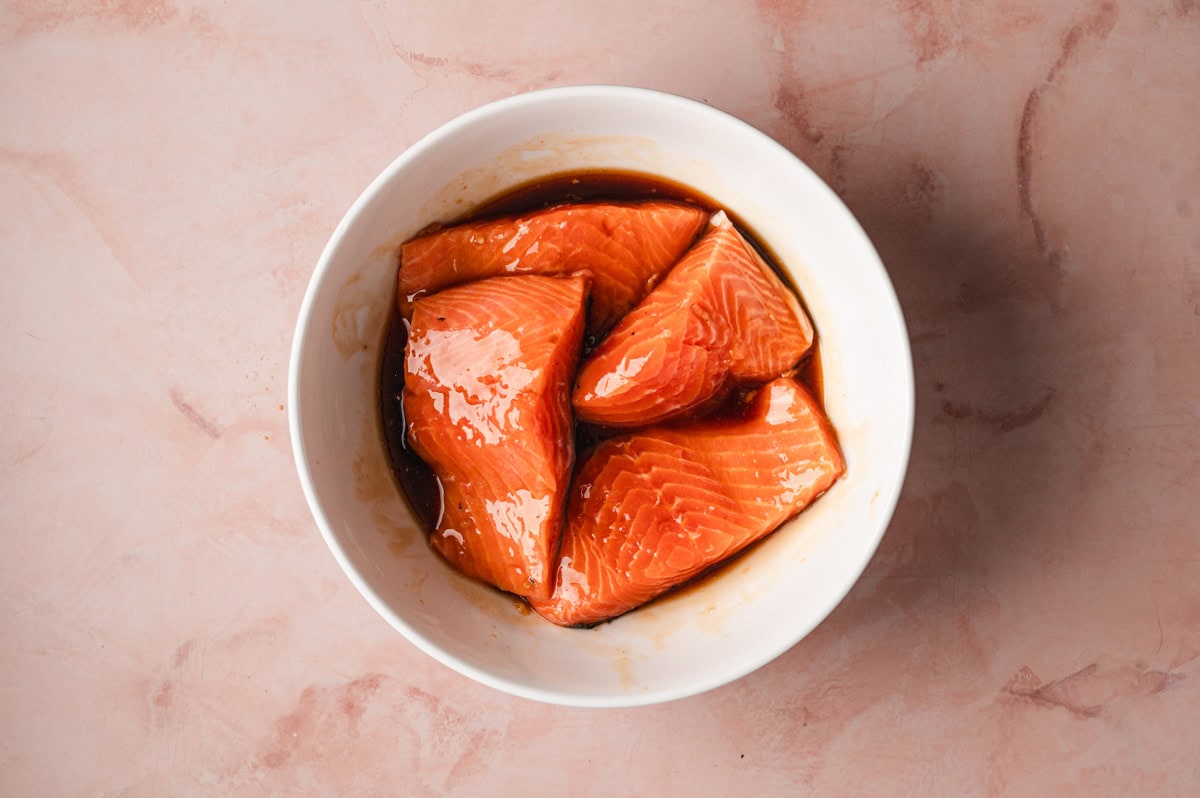 Four pieces of uncooked salmon in a teriyaki marinade in a white bowl.