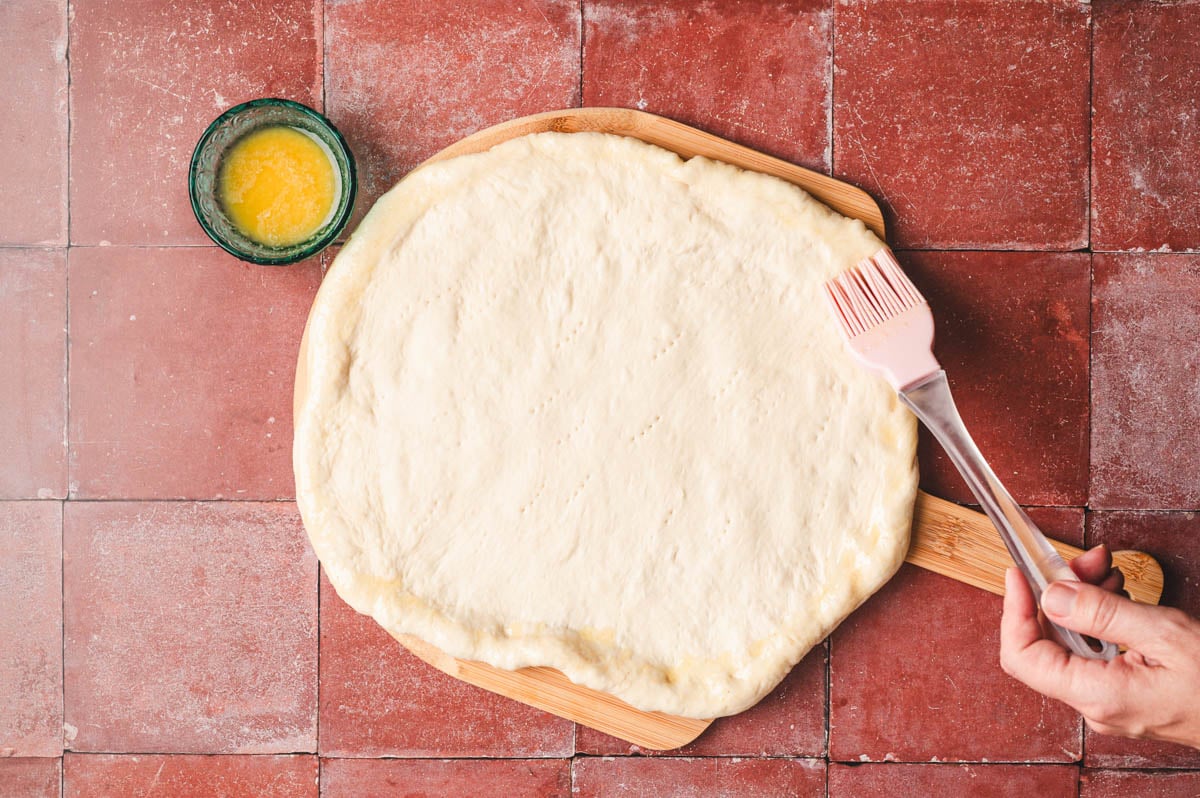 Pizza dough on a wooden board being brushed with butter.