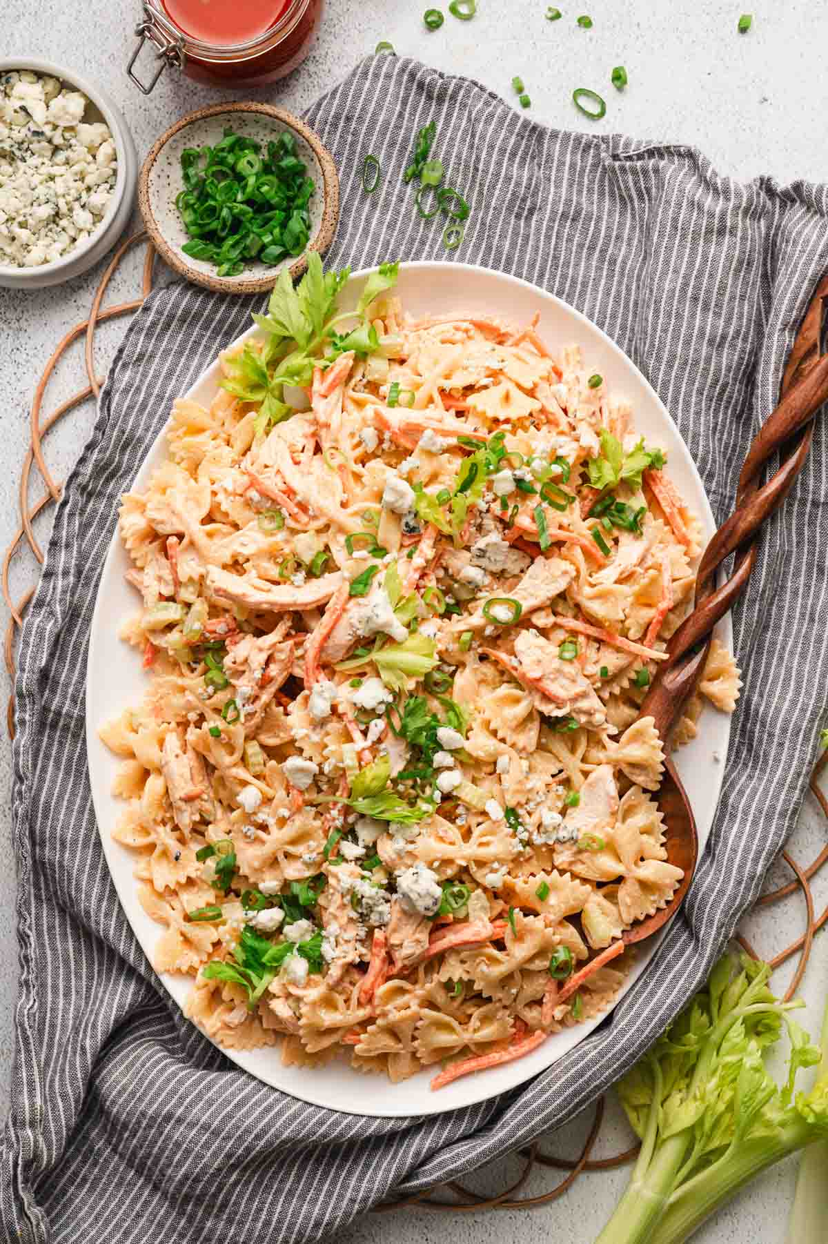 Buffalo chicken pasta salad in a large bowl pictured with sliced green onions, bleu cheese crumbles, and buffalo sauce.