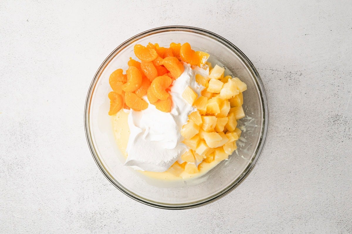 Glass mixing bowl with pudding, whipped topping, pineapple chunks, and mandarin oranges.