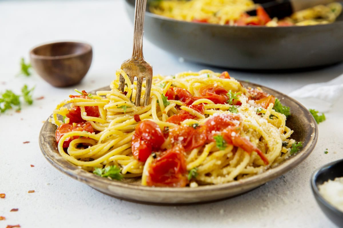 A plate of pasta with burst tomato and parsley, and a fork turning some noodles onto it.