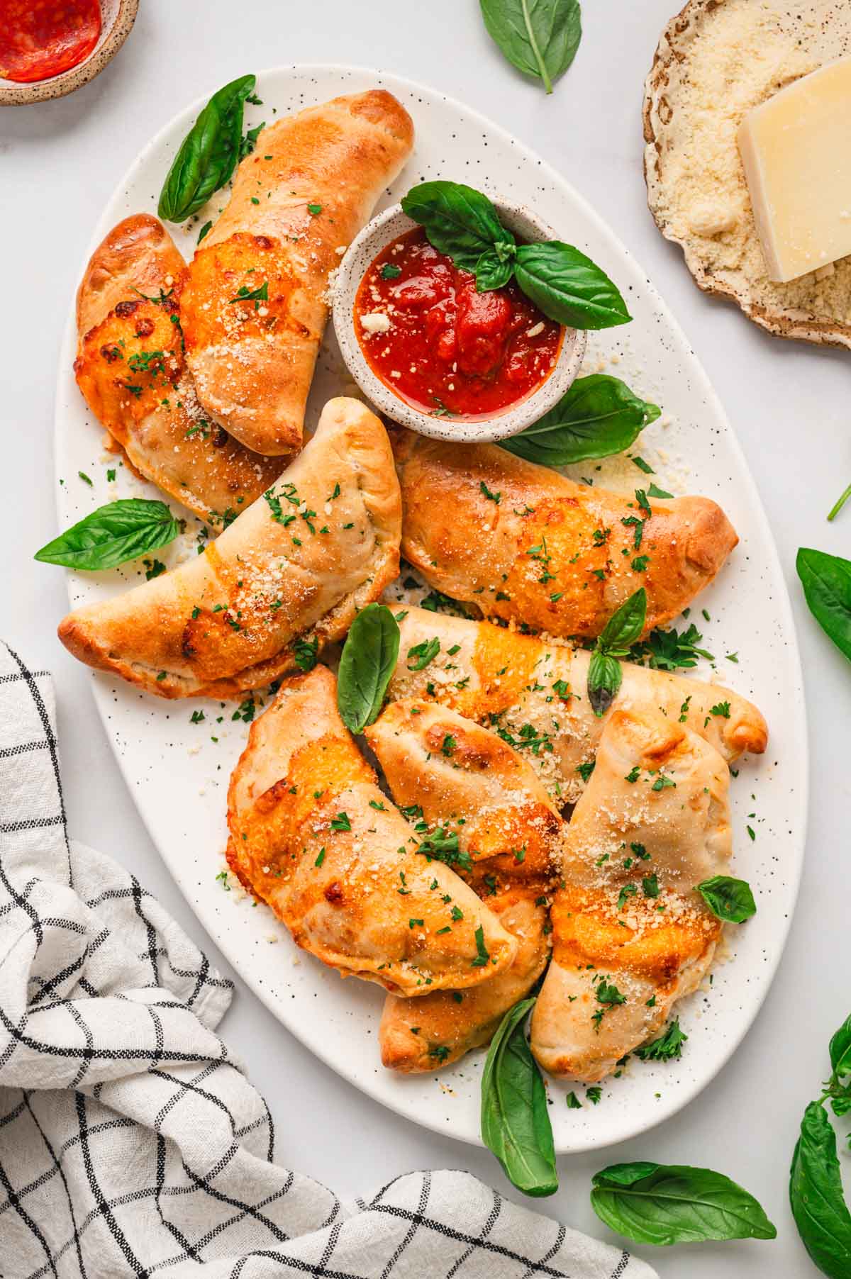 Baked mini calzones on a white serving platter with speckles on it. Garnished with fresh basil leaves.
