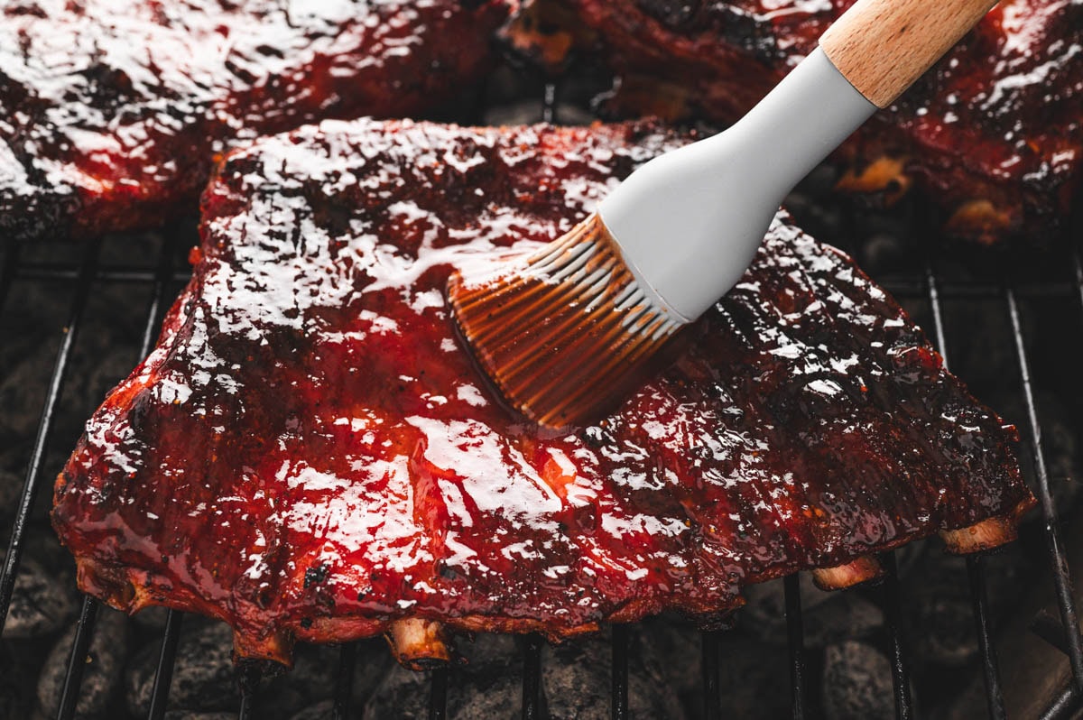 Cooked St. Louis ribs being brushed with BBQ sauce with a basting brush.