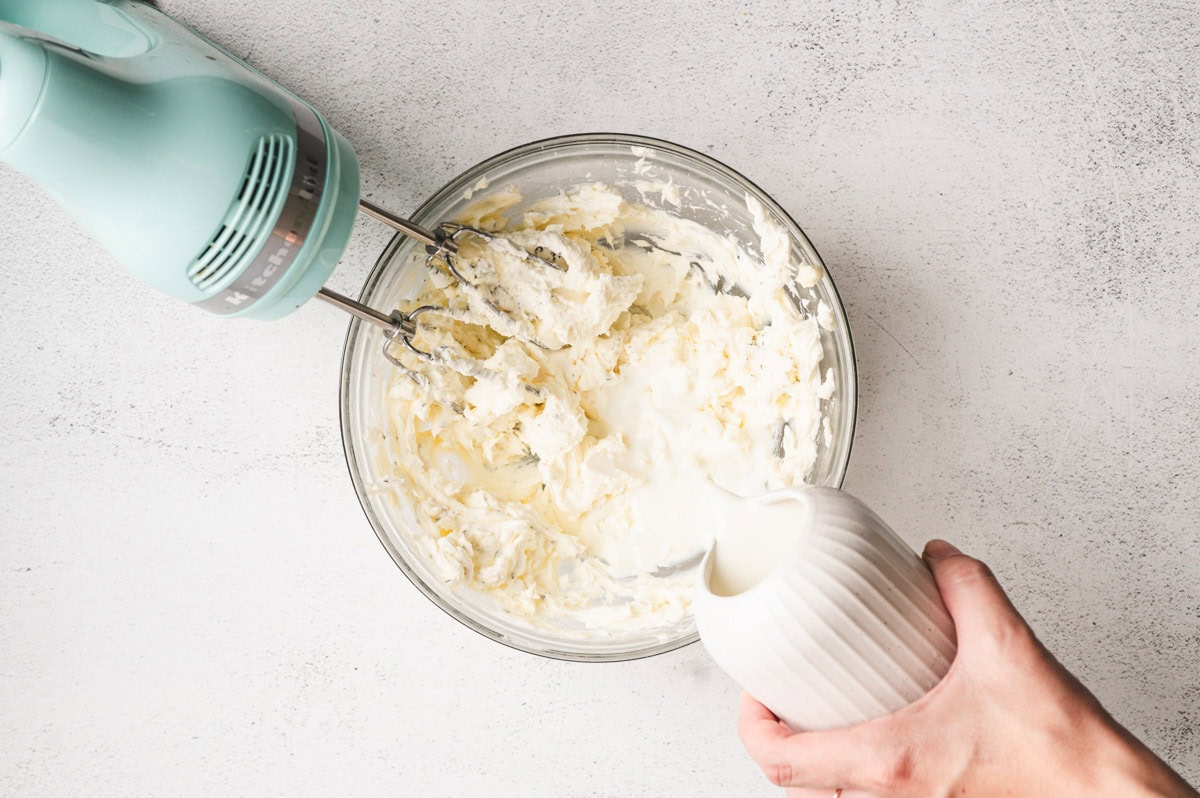 Cream cheese and butter in a glass bowl being mixed with a handheld mixer. Hand is pouring milk into the mixture.