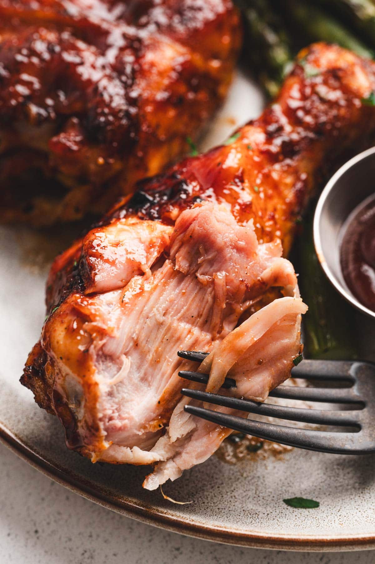 Close up of a fork and grilled BBQ chicken drumstick slathered in BBQ sauce on a plate.