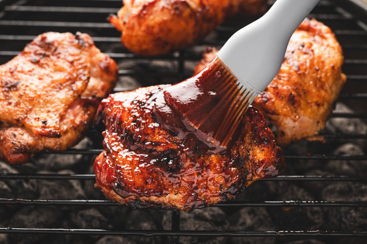 Skin on, bone in chicken thigh on a charcoal grill being basted in BBQ sauce with a brush.