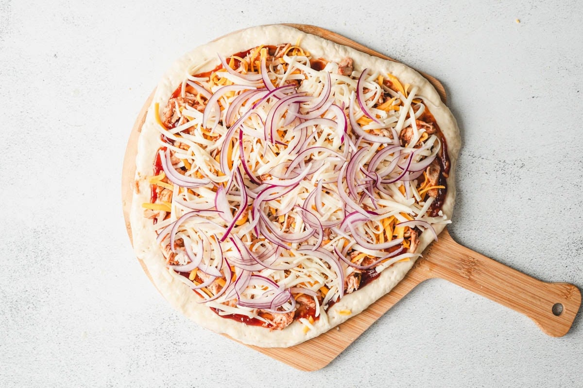 Uncooked pizza crust with BBQ sauce, chicken, cheese, and red onion on top.