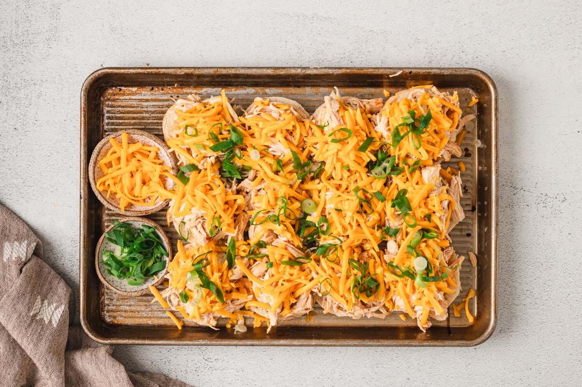 Bottom of slider buns on a sheet pan with shredded chicken, cheese, and sliced green onions on top .