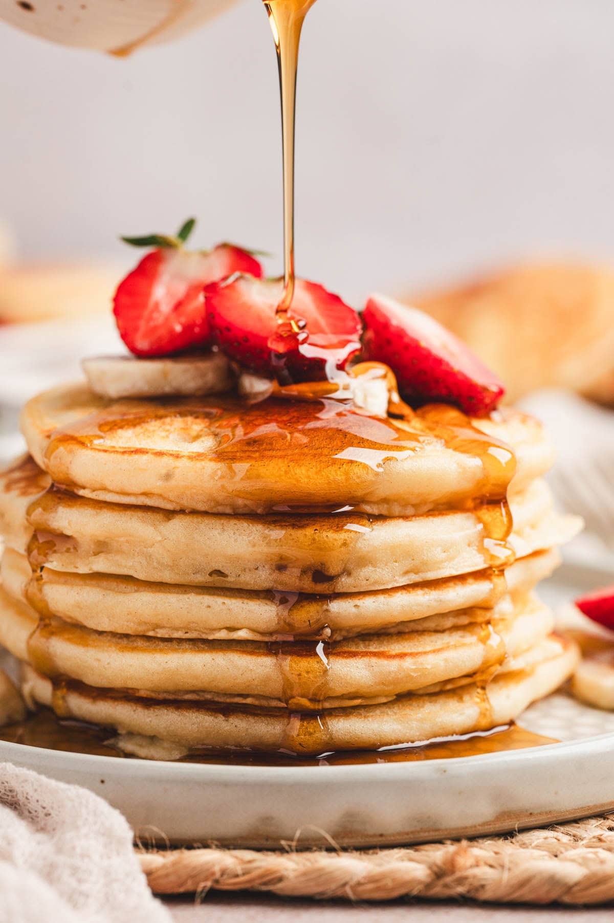 Buttermilk pancakes in a stack with fresh strawberries on top and maple syrup being drizzled on top.