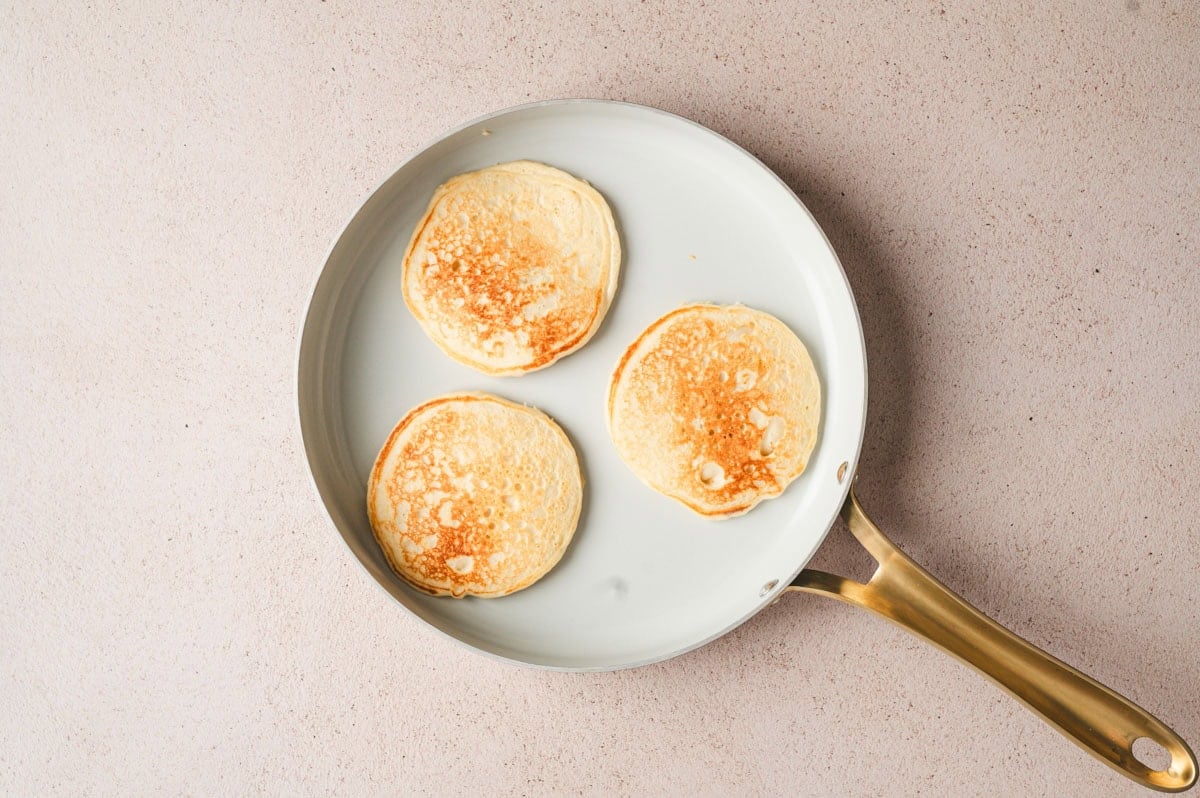 Cooked pancakes on a skillet after being flipped, showing one side with a golden-brown surface.
