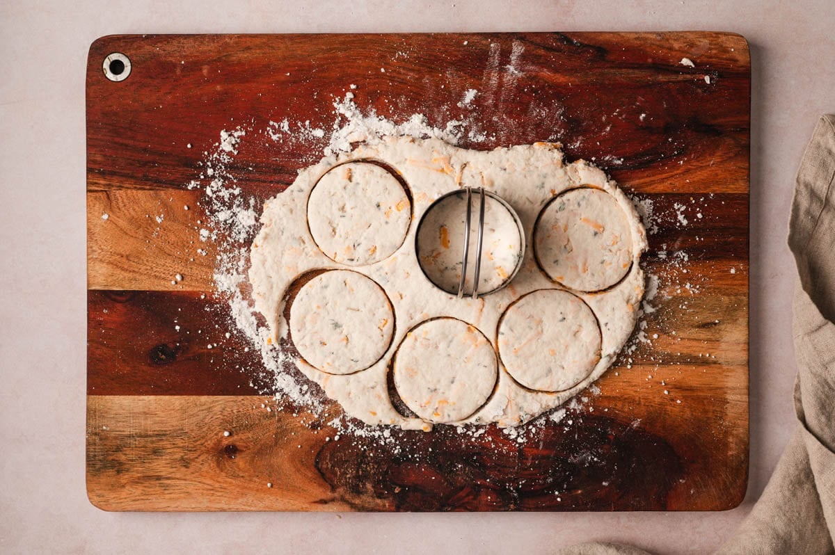 Biscuits being cut with a 4" biscuit cutter on a wooden butting board.