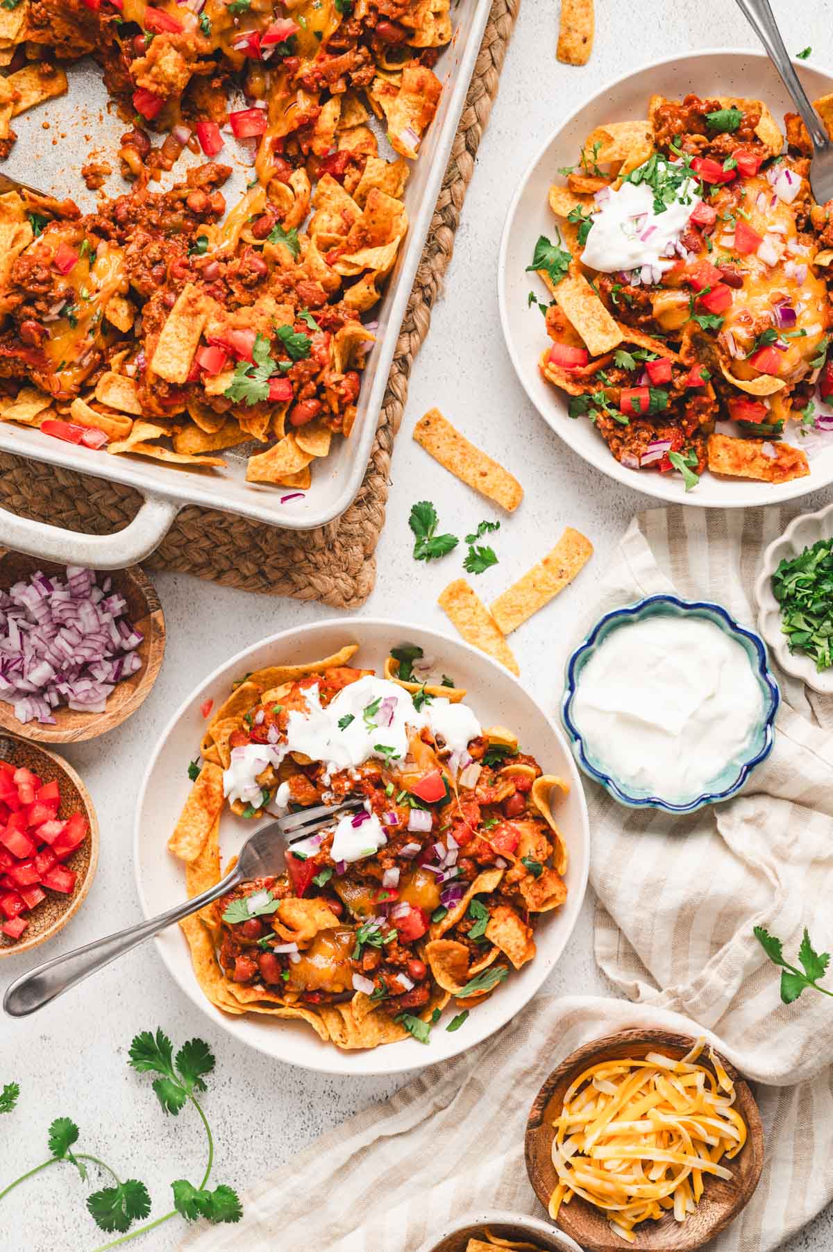 Plate with frito pie topped with onions, tomatoes, cilantro, and sour cream. Plate is sitting on a table with serving dish of frito pie and toppings.