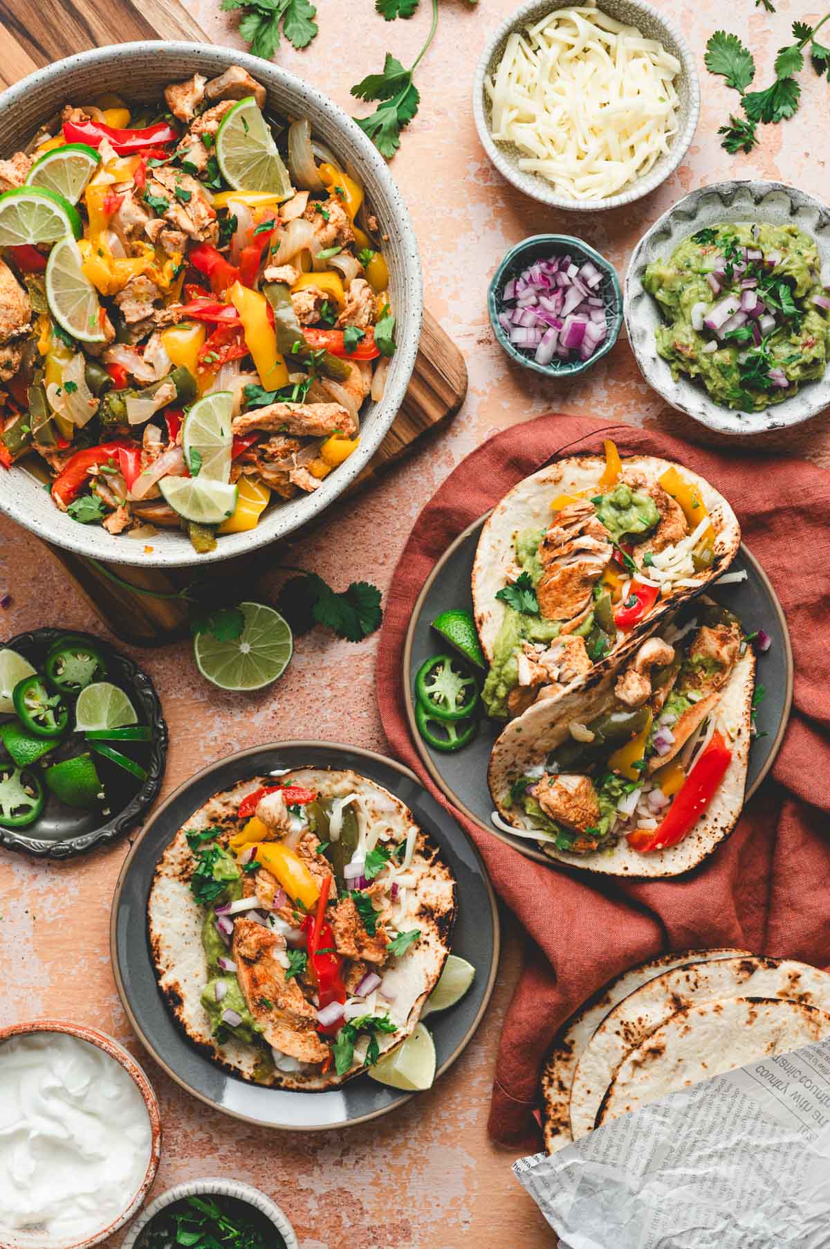 Plates of chicken fajitas with red onion, guacamole, cilantro, lime wedges, and jalapenos all set out on a table.