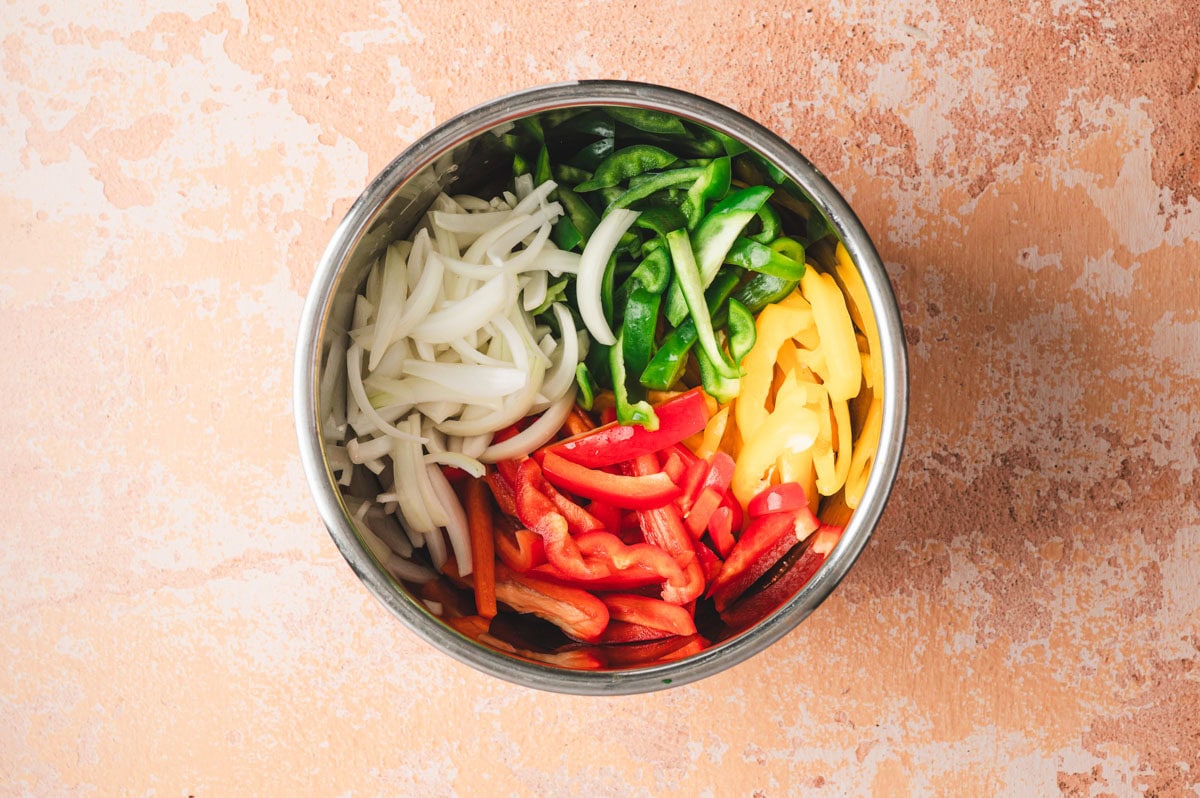 Sliced red, yellow, and green bell peppers and onions in a stainless steel pot.