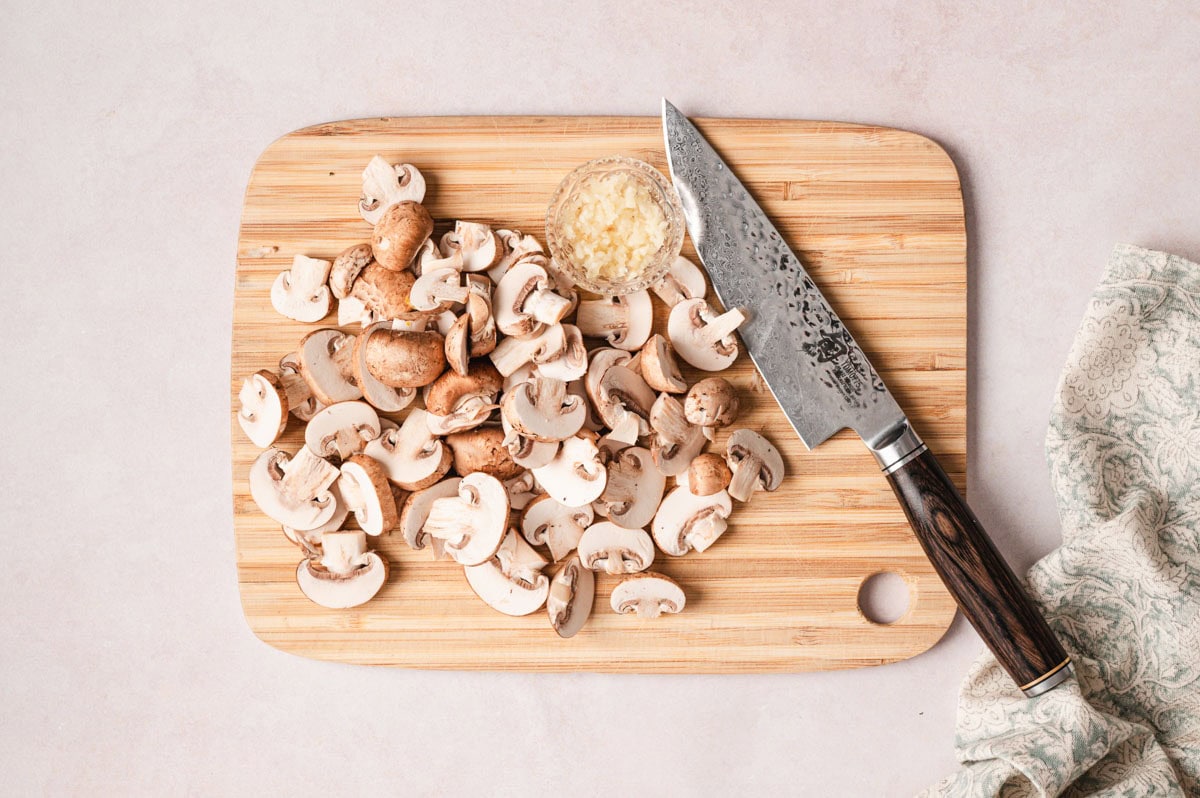 Mushrooms being chopped on a wooden cutting board. Minced garlic is in a clear glass dish next to the mushrooms.