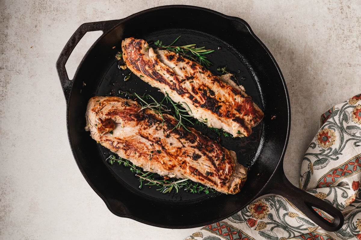 Cast iron skillet with two fully cooked pork tenderlloins with herbs.