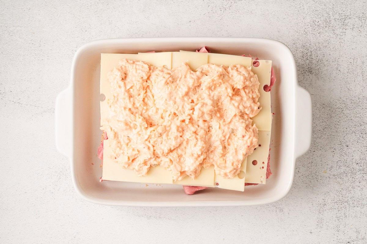 White casserole dish with slider buns topped with corned beef, swiss cheese, sauerkraut and thousand island dressing.