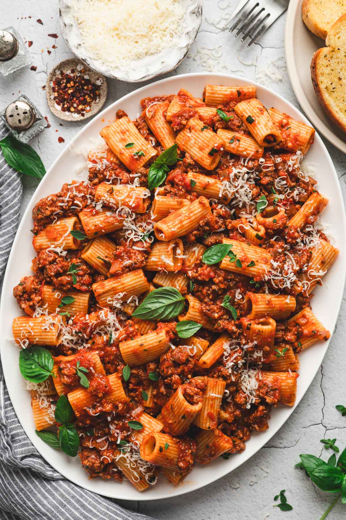 Rigatoni with bolognese sauce on a white platter, topped with parmesan and fresh basil leaves.