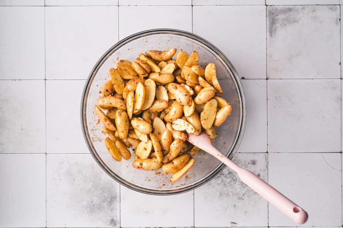 Uncooked sliced fingerling potatoes in a glass bowl tossed with olive oil and seasonings.