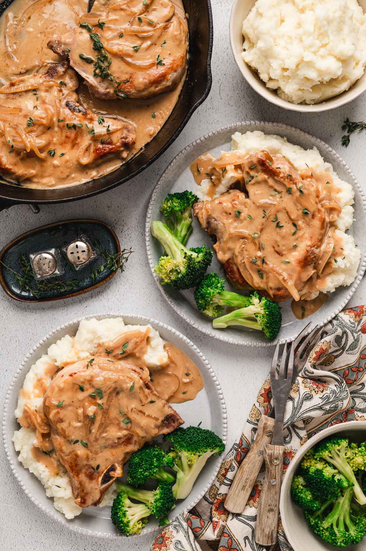 Smothered Pork Chops with creamy sauce and onions over mashed potatoes with broccoli, shown on two grey dinner plates.