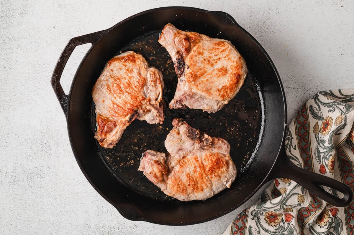 Pork chops being seared in a cast-iron skillet.