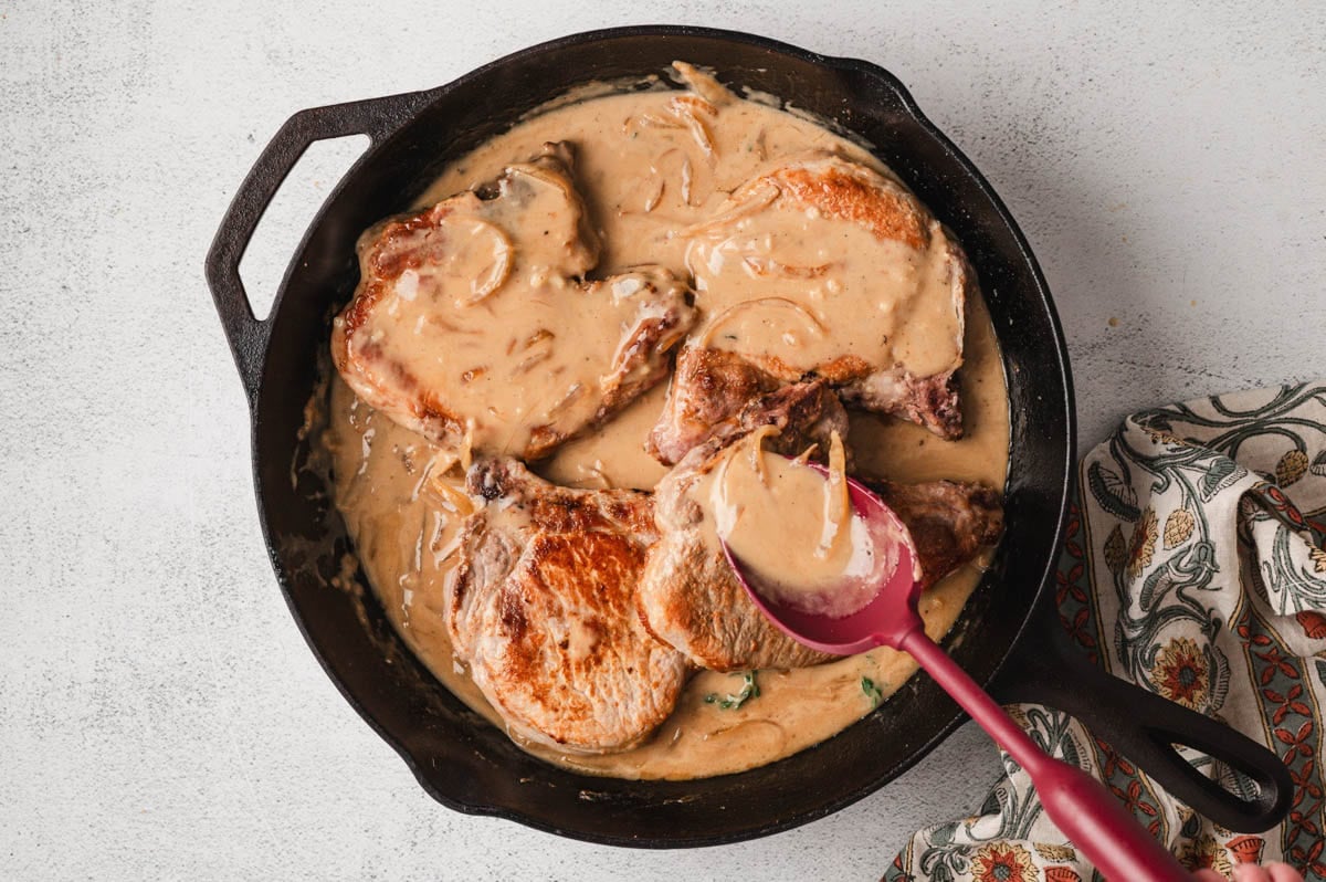 Cooked smothered pork chops in a cast-iron skillet with a red serving spoon in the skillet.