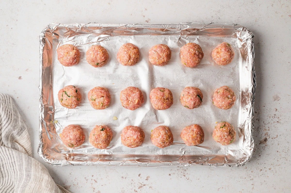 Raw meatballs lined up on a foil lined baking sheet.