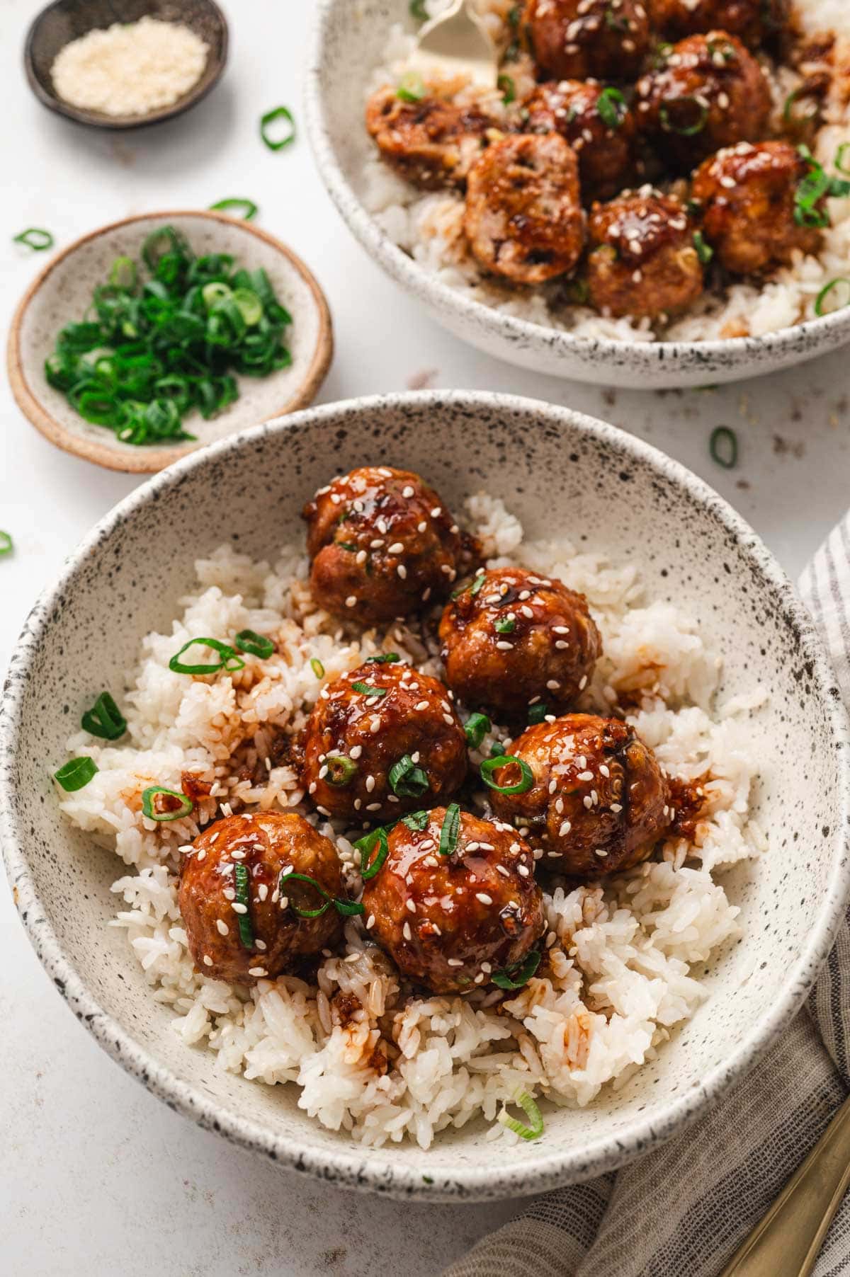 Teriyaki meatballs served in a bowl over white rice. Topped with green onions and sesame seeds.