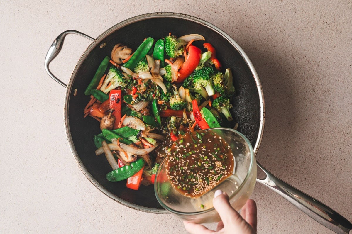 Stir fry sauce being poured over the vegetables in a large skillet.
