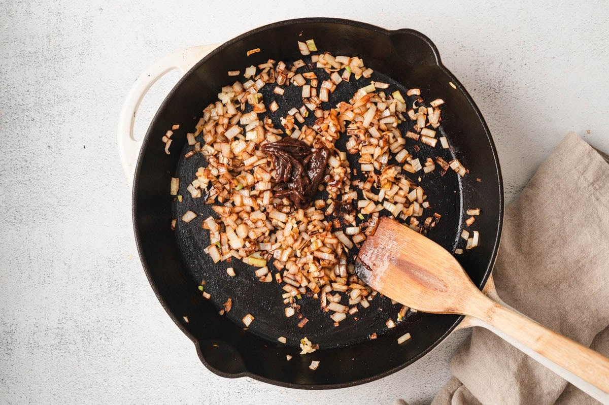 Onion and garlic being sauteed in a cast iron skillet with a dollop of beef base (bouillon concentrate) and a wooden spatula.
