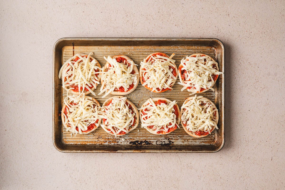 English muffins on a sheet pan topped with red sauce and cheese.