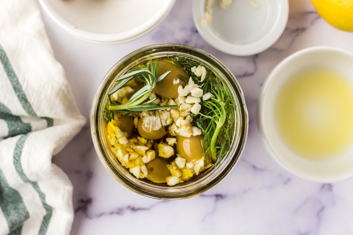Overhead image of a small jar filled with olives, olive oil, garlic and herbs.