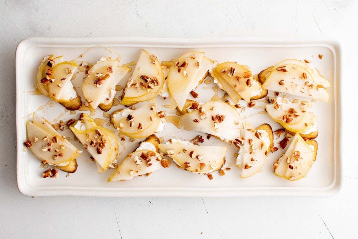 Pear and goat cheese crostini with honey and pecans on a white platter.