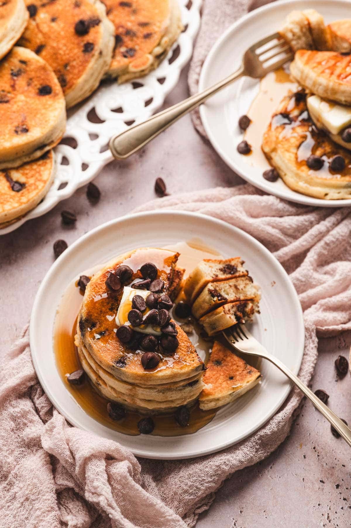 Plate with stack of chocolate chip pancakes topped with butter, maple syrup, and chocolate chips, with a fork cutting into them.