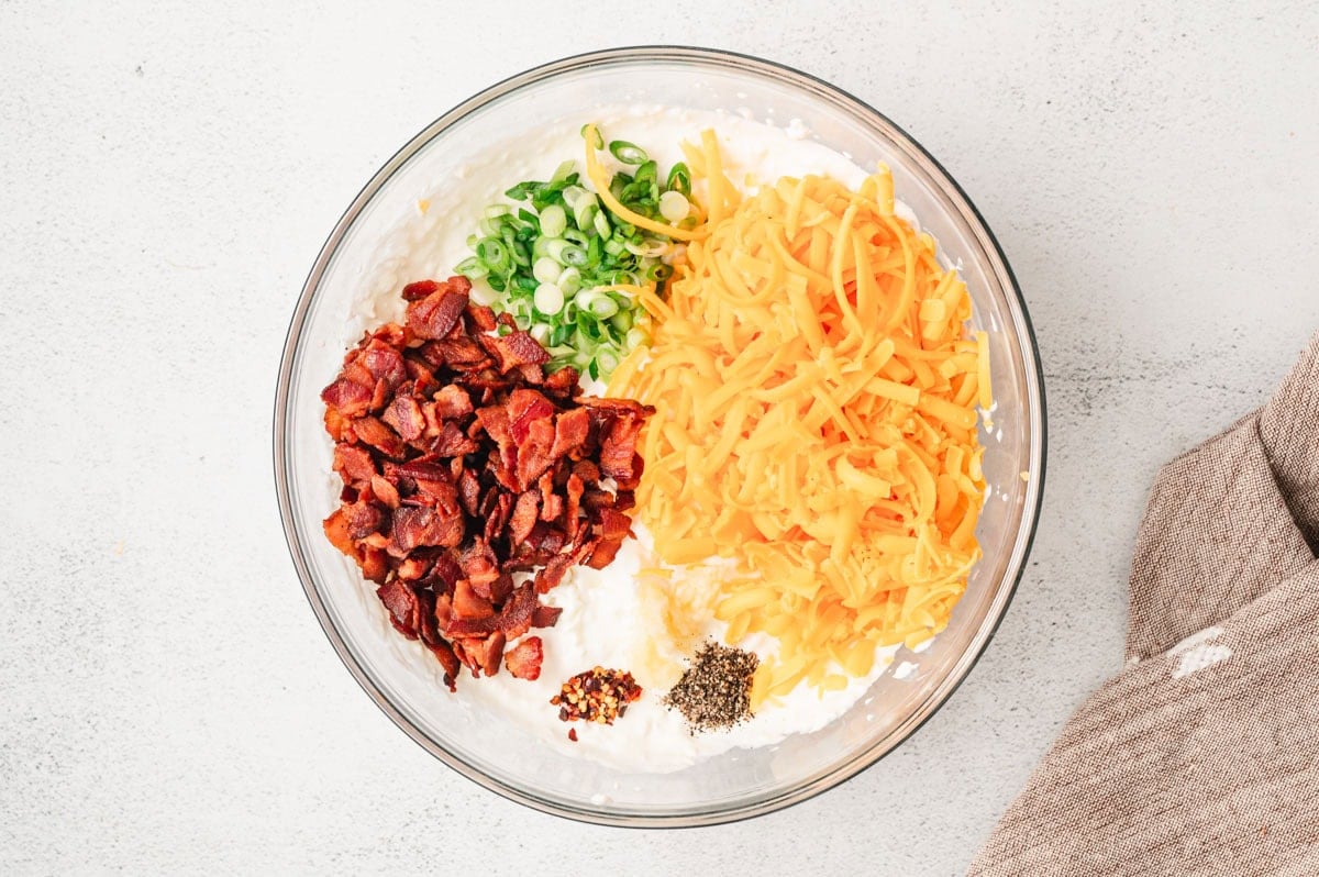 Ingredients for hot bacon dip in a glass bowl - cream cheese, sour cream, mayo, seasonings, crispy bacon pieces, shredded cheddar cheese and sliced green onion.