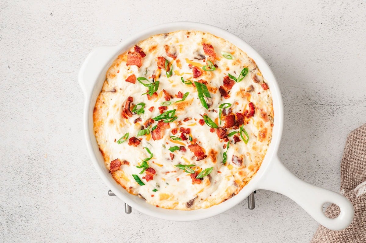 Fully baked hot bacon dip in a white dish, topped with bacon, green onions.