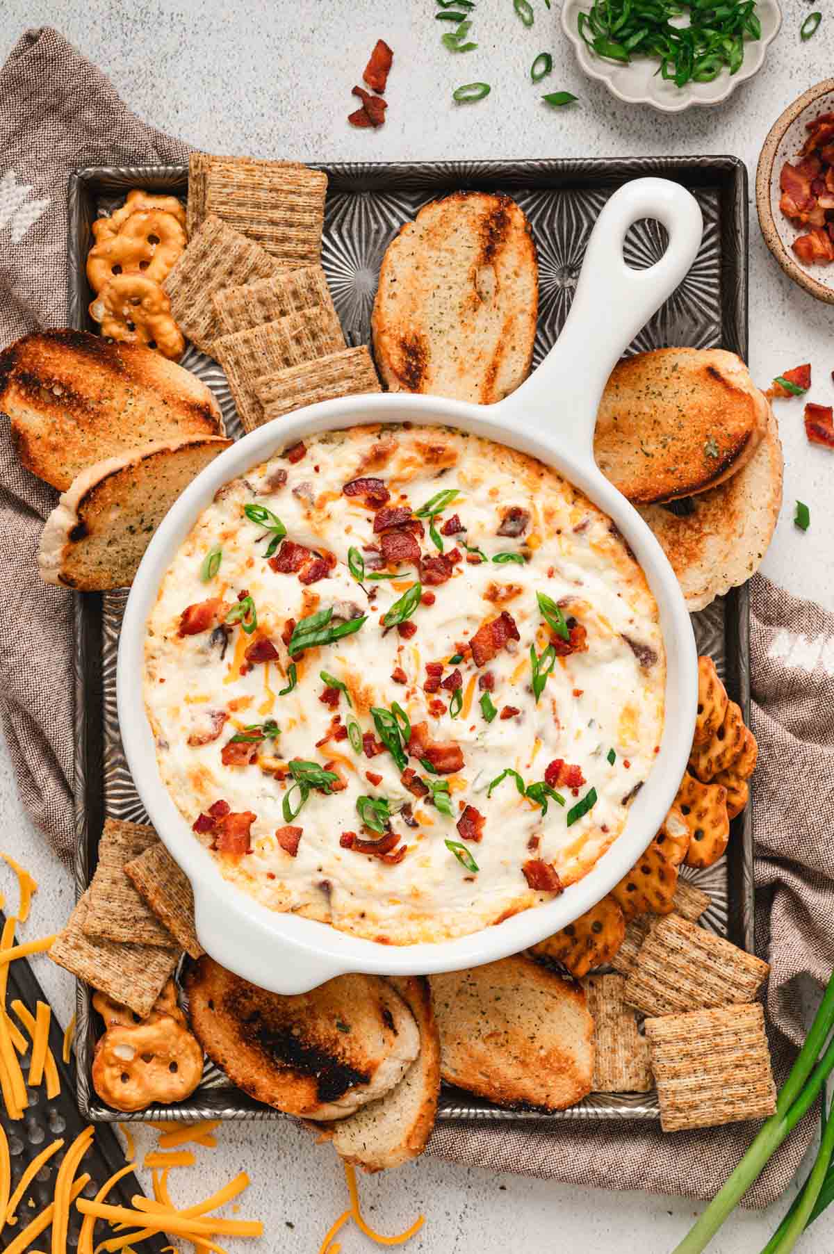 White dish with hot bacon dip garnished with bacon, green onions on top, on a sheet pan with crackers, pretzels, and crostini.