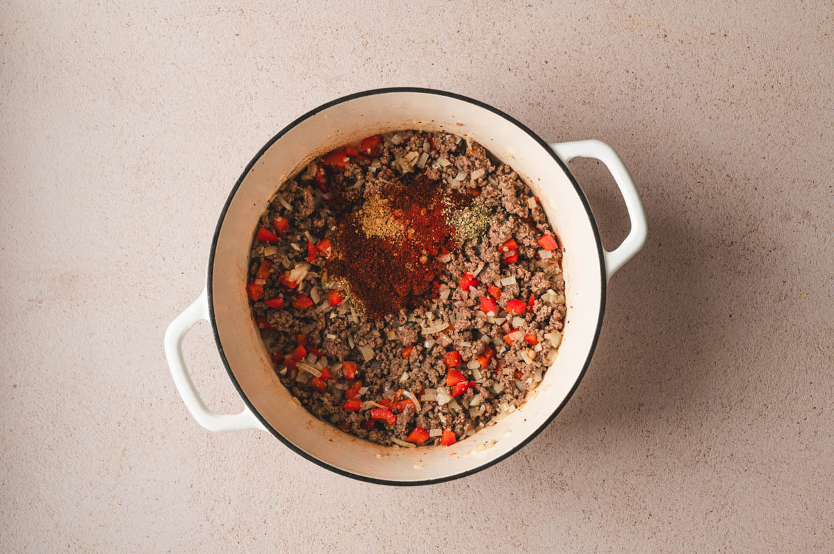White dutch oven with ground beef, diced red peppers and onions and spices.
