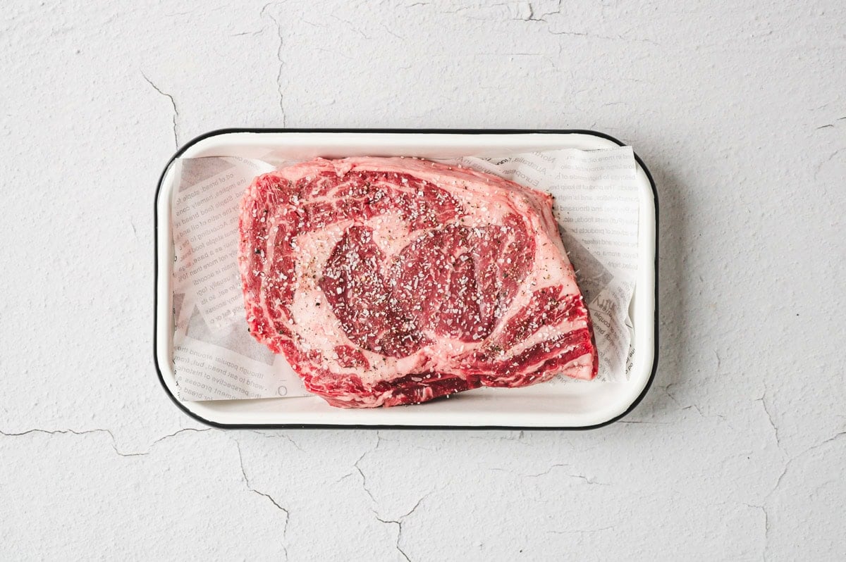 Uncooked ribeye steak on a tray with seasoning on top.